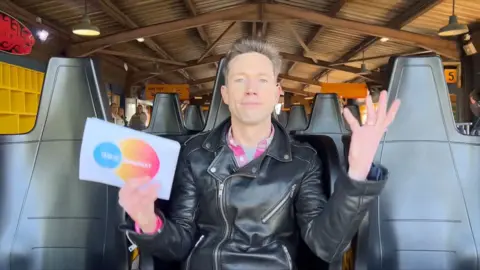 Peter Ruddick is sat on a roller coaster wearing a black leather jacket. He is holding a white cue card.