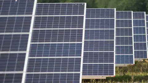 PA Media A number of solar panels in a field. There is a number of blue panels with a silver pattern.