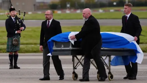 PA Media Alex Salmond's coffin draped in a saltire at Aberdeen airport, with a bagpiper in the background