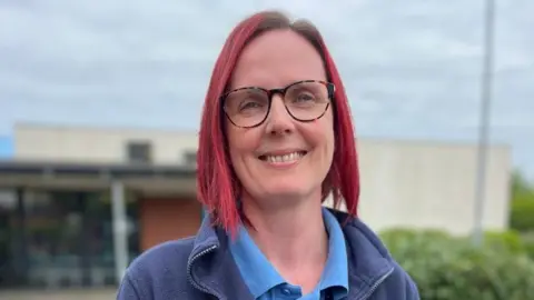 A woman with died red hair in a bob, and glasses, is smiling at the camera. She is wearing a blue district nurse polo shirt with a purple lanyard and blue fleece over the top. The NHS logo is visible on her left hand breast pocket.