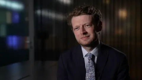 Connor Rand smiles, wearing a black suit, white shirt and floral tie, as he is interviewed in an office room