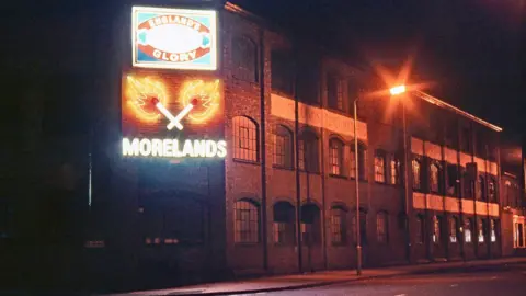 A photo of a red bricked factory with a lit up England's Glory sign and a lit matches and Moreland's sign