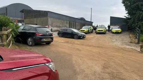 BBC Cars and police vehicles sit on a dirt path near farm buildings