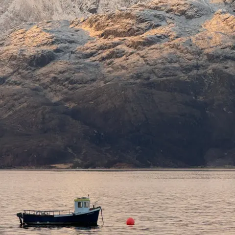 Heather McLean A small fishing boat floats on calm water in the foreground, with a red buoy nearby. Snow-dusted mountains rise steeply behind, their upper slopes catching warm light under a soft, overcast sky.