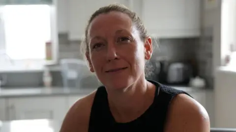 John Fairhall/BBC Tracy Williams is wearing a black vest while sitting in a kitchen. She has her light brown hair in a ponytail.