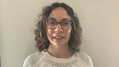 a woman with brown curly hair and glasses, wearing a white top