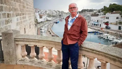 Swansea Bay University Health Board David is stood on the balcony of a stone building, overlooking a harbour where yachts and small boats are moored. There are rows of white buildings in the background. He is wearing a blue and white patterned shirt and a brown jacket and blue trousers, leaning against the balcony wall, with his hands in his pockets.