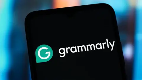 Getty Images A close-up of a smartphone screen displaying the white and green Grammarly logo against a black background. The phone is held against a soft-focus, blue and orange bokeh background.