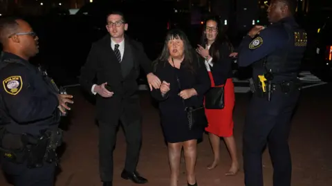 Niall Carson/PA Wire Enoch Burke's brother, mother and sister remonstrating after being ejected from the National Building Museum.  Mrs Burke is wearing a black dress and shoulder bag but has lost one of her shoes.  Ammi is wearing a red shirt and a black jacket and she has lost both of her shoes. 