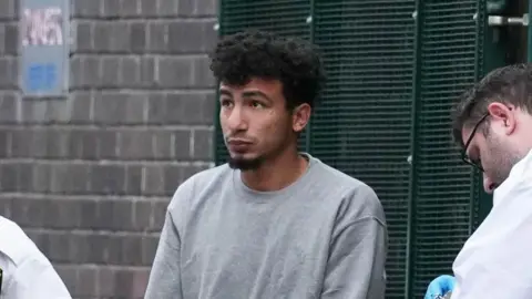 A man with black hair, wearing a grey jumper. He is standing between two custody officers. 