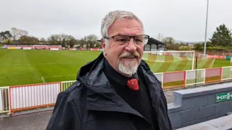 Lester standing in front of the Frome football pitch wearing glasses and a red tie