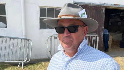 Farmer Mark Wagstaff stands in front of a cow shed at the Great Yorkshire Show. He's wearing a brown hat and a blue check shirt.