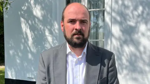 Richard Holden a man standing outside a white building. He is wearing a grey suit jacket and white shirt. He is looking directly at the camera.