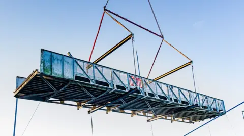 East Riding of Yorkshire Council A metal bridge being lifted by crane cables. The grey structure is suspended in the air against a clear blue sky.