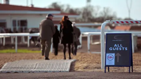 PA Media Smartly-dressed visitors to the racecourse walking towards the entrance of a white building with an orange tiled roof. There is a sign in the foreground that shows which way all guests need to go.
