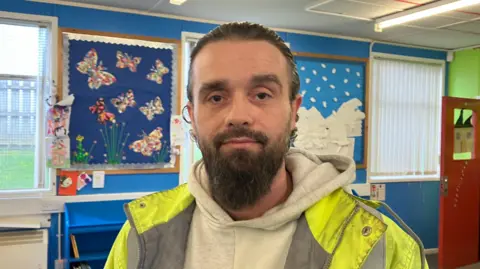 Arron Cooley has slicked back black hair and a beard. He is standing in a classroom. He is wearing a white hoodie underneath a fluorescent safety jacket.