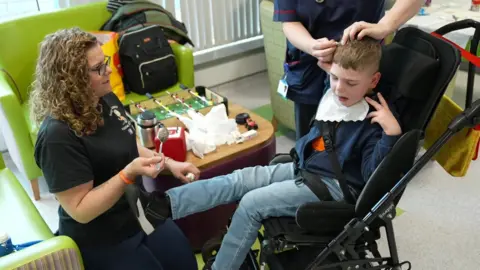 Aimee Tilley on her knees holding a spoon, wearing a black T-shirt looking at her son Isaac who is in a wheelchair in a hospital setting