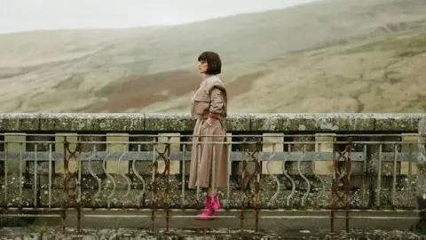 Indie-Lincs Film Festival A screengrab from Wild Animal. It shows a woman with a short brown bob wearing a beige outfit and bright pink boots standing on a bridge footpath