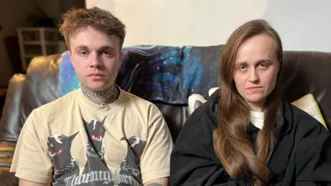 A man and a woman sit together on a sofa facing the camera. The man has a cream T-shirt on with black dogs on it. The woman has long dark brown hair and is wearing a black top