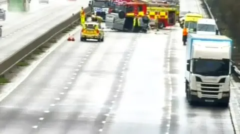 A closed four-lane stretch of road. In the middle is an overturned silver van. There are emergency services including fire, ambulance and police vehicles.