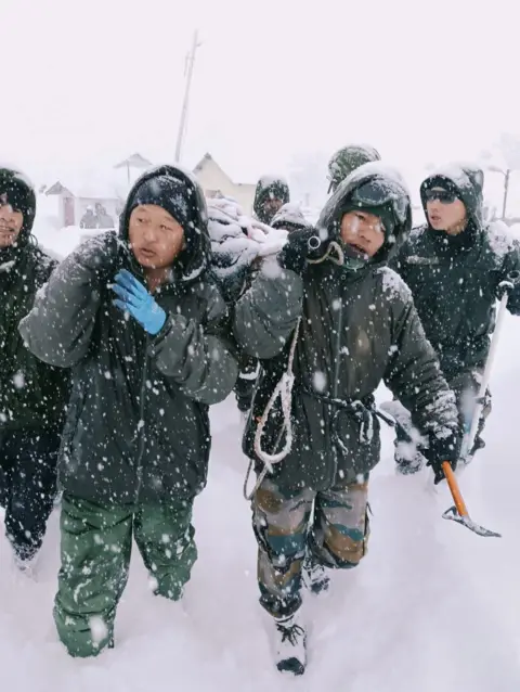 Two rescuers from the Indian army and two rescuers from the Indian army, are wearing the green overalls, carrying items over their items when they walked with heavy snow