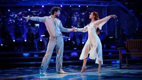 George Clarke and his dance partner Alexis Warr on Strictly Come Dancing in 2025. They are stood holding hands at an arm's length apart, holding their arms out behind them.