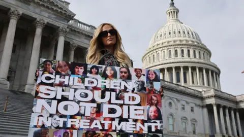 Getty Images Celebrity hotel heiress Paris Hilton poses for photographs outside the US Capitol in December 2024 carrying a poster that says "Children should not die in treatment".