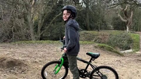 A boy called Rex is seen standing with a bike under him - he has a crash helmet on. He is in a wooded area