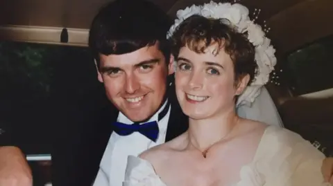 Fulton family PC Lewis Fulton and wife Christine on their wedding day. PC Fulton is wearing a dark suit with a white shirt and blue bow tie. Christine is wearing a white dress and white floral headband with a veil at the back.