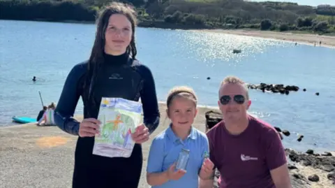 Chloe, Eliza and Matt are standing on a beach on a sunny day and there is a small bay behind them. Chloe is holding a picture drawn by Leo that was in the jam jar that Eliza is holding. Eliza is also holding the Lego spaceman. They are all smiling. 