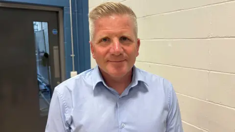 Chris Quinn, a man with short blond hair, smiles at the camera. He is wearing a light-blue shirt and is standing next to a white wall. Behind him is a door. 