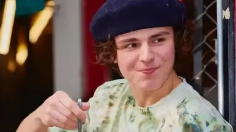 Family photo He is wearing a blue flat cap and has bushy, brown curly hair underneath. He is looking to the right and appears to be holding a piece of cutlery. He is wearing a green patterned T-shirt and leaning up against a wire fence.