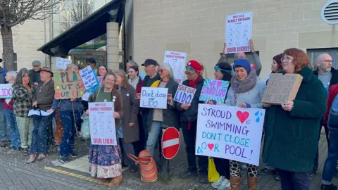 Carmelo Garcia A row of around 20 people holding placards outside Ebley Mill, where councillors are discussing the future of the lido. The signs say things like 'Stroud loves swimming, don't close our pool', and 'we love Stroud lido'.