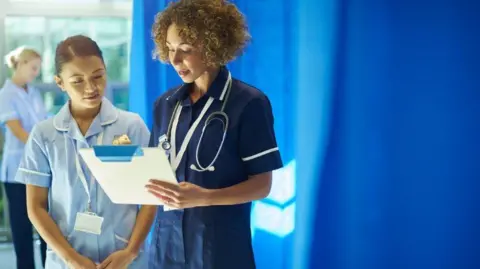 A chief nurse stands with a student nurse explaining the contents of a chart to her. They are wearing blue nurses' uniforms and there is a blue curtain behind them.