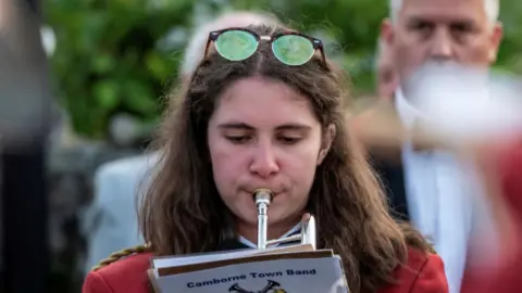 Paul Williams A woman called Lia Teague. She is 22. She is playing a cornet and has glasses propped on her head. She has shoulder length brown hair. Bandmates are stood behind her.