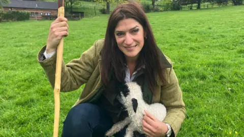 A woman with long brown hair, a green jacket and jeans, smiles as she kneels down in a field while holding a lamb in her left arm and holding a shepherdess's cane in her right hand.