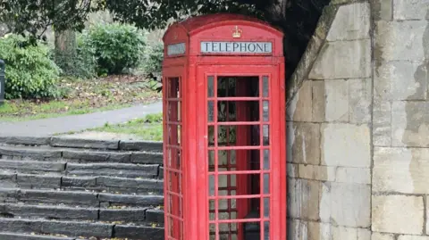 Bath Preservation Trust A red phone box without its windows is in the foreground. Behind it on the right is a wall, and on the left are some steps leading to a wooded footpath.