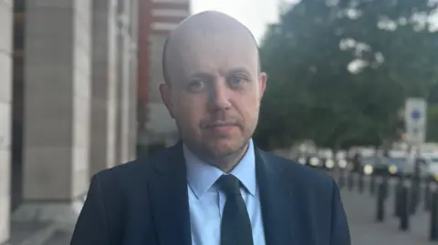 A bald man in a blue suit, light blue shirt and dark tie stands on a London street. He looks into the camera.