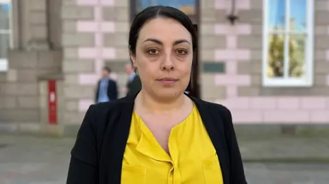 Deputy Carina Alves - A woman wearing yellow shirt standing in front of states building 