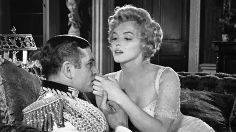 Park Circus A scene from The Prince and the Showgirl film, in black and white. Marilyn Monroe leans next to a man dressed as a prince, in formal attire. 