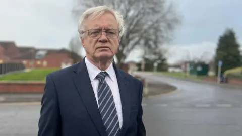 David Davis standing facing the camera. He has grey short hair. He is wearing glasses, a navy suit and a dark blue and light blue tie. A road and residential houses are in the background. 