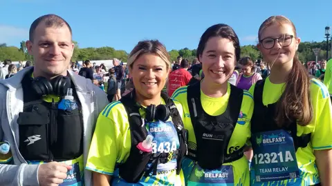 There are four people in the picture. From left to right is a dark haired man with a grey jacket and black running vest. There are three women next to him with a hi-vis top on and black running vest. 