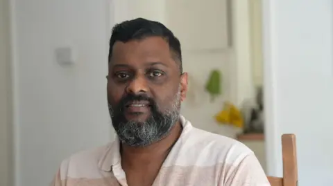 Sumesh has a dark beard and is wearing a beige polo shirt. He is sitting in a wooden chair and smiling at the camera