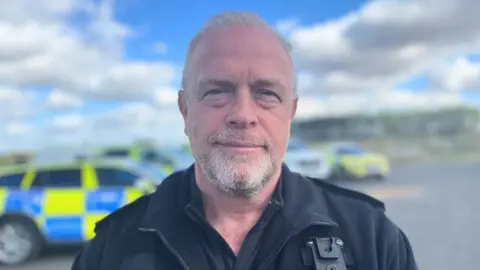 Insp Chris Davis, who has grey hair and a neatly trimmed beard, wears a black police uniform and stands in front of marked police cars.