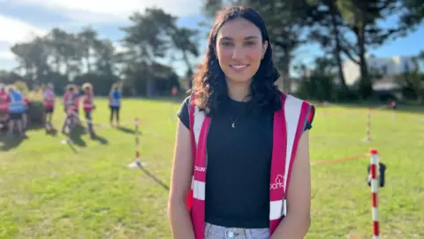 BBC Saesha smiles at the camera wearing a highviz Parkrun vest as volunteers get ready behind her