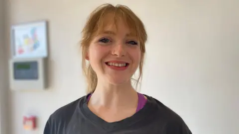 A woman with red hair tied up in a ponytail is wearing a black T-shirt. She is in front of a white wall.