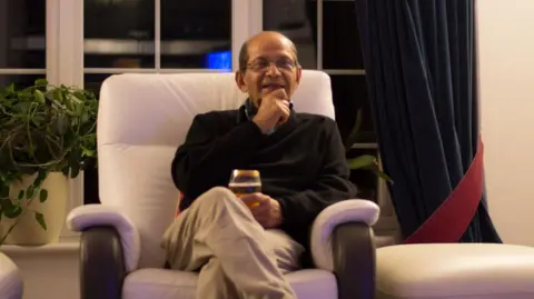An elderly man sits in a chair and smiles. He looks like he might be speaking. He holds a class in his lap and sits upright in a navy sweater.