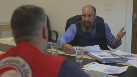 Two men sit apart across a large conference room desk, which is strewn with papers, a laptop and glasses of water. One man, with dark greying hair, sits with his back to the camera and is wearing an out of focus red vest on top of his navy shirt. The other man is dressed in a a shirt, tie and waistcoat. He has dark greying hair and a long beard. His hands in the air while holding documents.