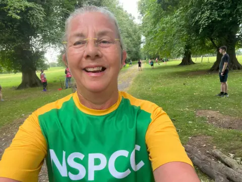 Sheila Mitchell A woman is smiling at the camera. She is wearing a green and yellow top that reads "NSPCC" and glasses. She is stood in a park with some large trees and a few people stood behind her in the distance. 