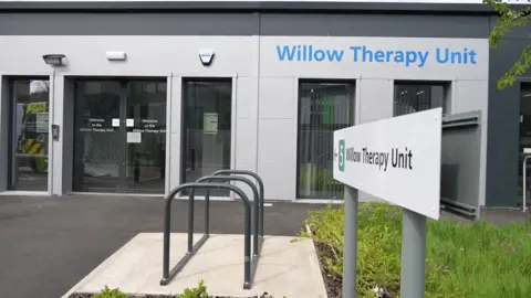 Qays Najm/BBC The Willow Therapy Unit has a sign outside of it, next to a small area of grass and a bike rack, which says its name. The grey building has an entrance on the left and several windows.